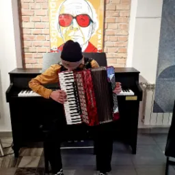 Młody mężczyzna w czapce gra na akordeonie marki Weltmeister, siedząc przed pianinem; w tle plakat z wizerunkiem osoby w czerwonych okularach na ceglanej ścianie.