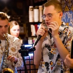 Muzyka - Młodzi muzycy w kwiecistych koszulach: saksofonista, wokalista w okularach z mikrofonem i gitarzysta podczas występu na tle regałów z książkami i roślinami.