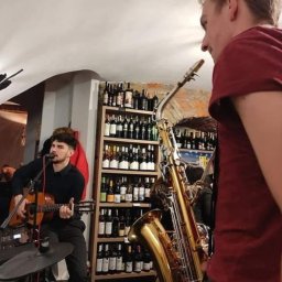 Muzyka - Występujący muzyk grający na gitarze i śpiewający do mikrofonu, obok saksofon, półki z butelkami w tle.