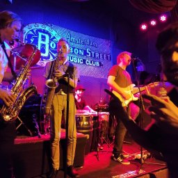 Muzyka - Zespół muzyczny grający na scenie w klubie Bon Street Amsterdam, oświetlenie sceniczne w kolorach niebieskim i czerwonym, saksofony, gitara elektryczna i perkusja.