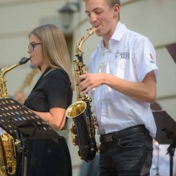 Muzyka - Dwoje młodych ludzi gra na saksofonach podczas występu plenerowego; mężczyzna w białej koszuli z logo orkiestry dętej.