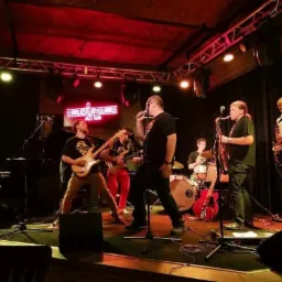 Zespół muzyczny grający jazz na scenie klubu z czerwonym oświetleniem, widoczni: wokalista, dwóch saksofonistów, gitarzysta basowy, gitarzysta, perkusista i pianista.