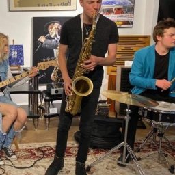 Muzyka - Saksofonista w czarnym ubraniu gra na saksofonie na tle zespołu muzycznego w pomieszczeniu z plakatami na ścianach i wzorzystym dywanem.