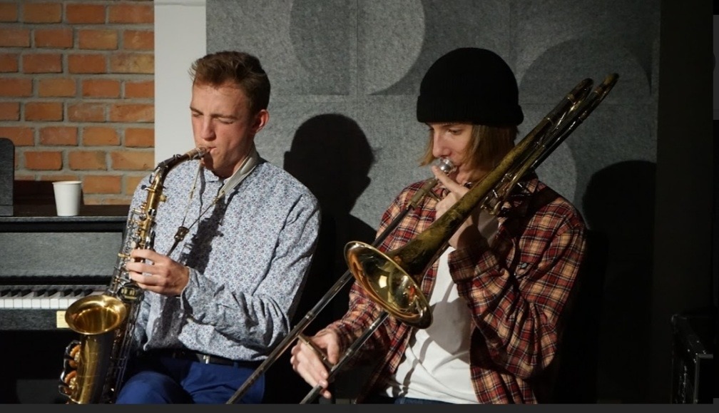 Duet muzyczny: saksofonista w kwiecistej koszuli i puzonista w czapce grający na tle ceglanej ściany i szarego tła z okręgami.