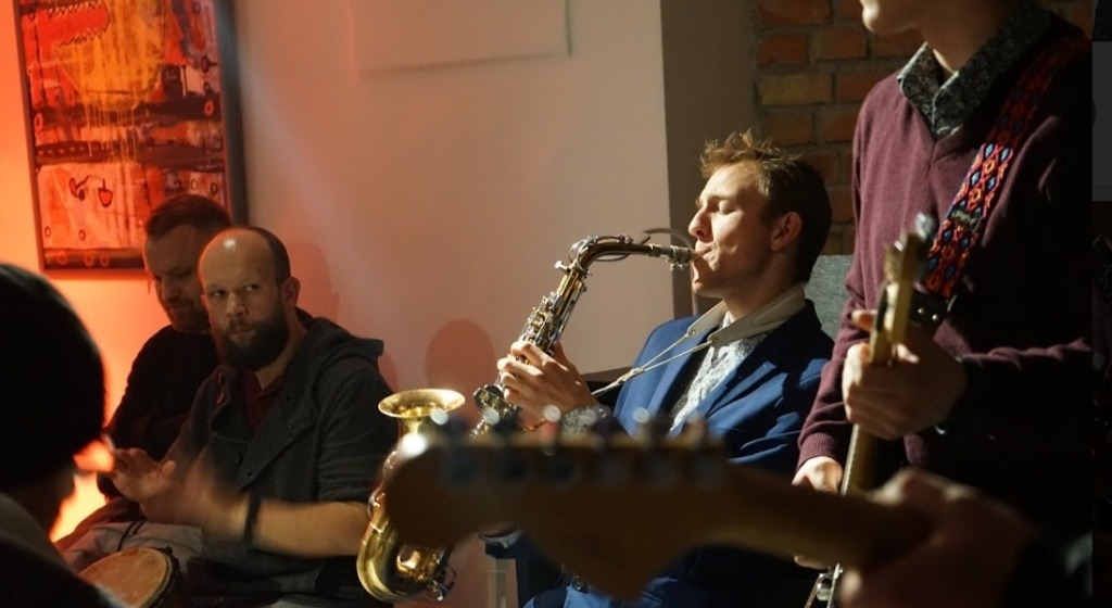 Występ zespołu w Bielsku-Białej: saksofonista w niebieskiej marynarce gra na saksofonie, obok gitarzysta i perkusista z bębnem djembe, w tle obraz w ciepłych barwach.