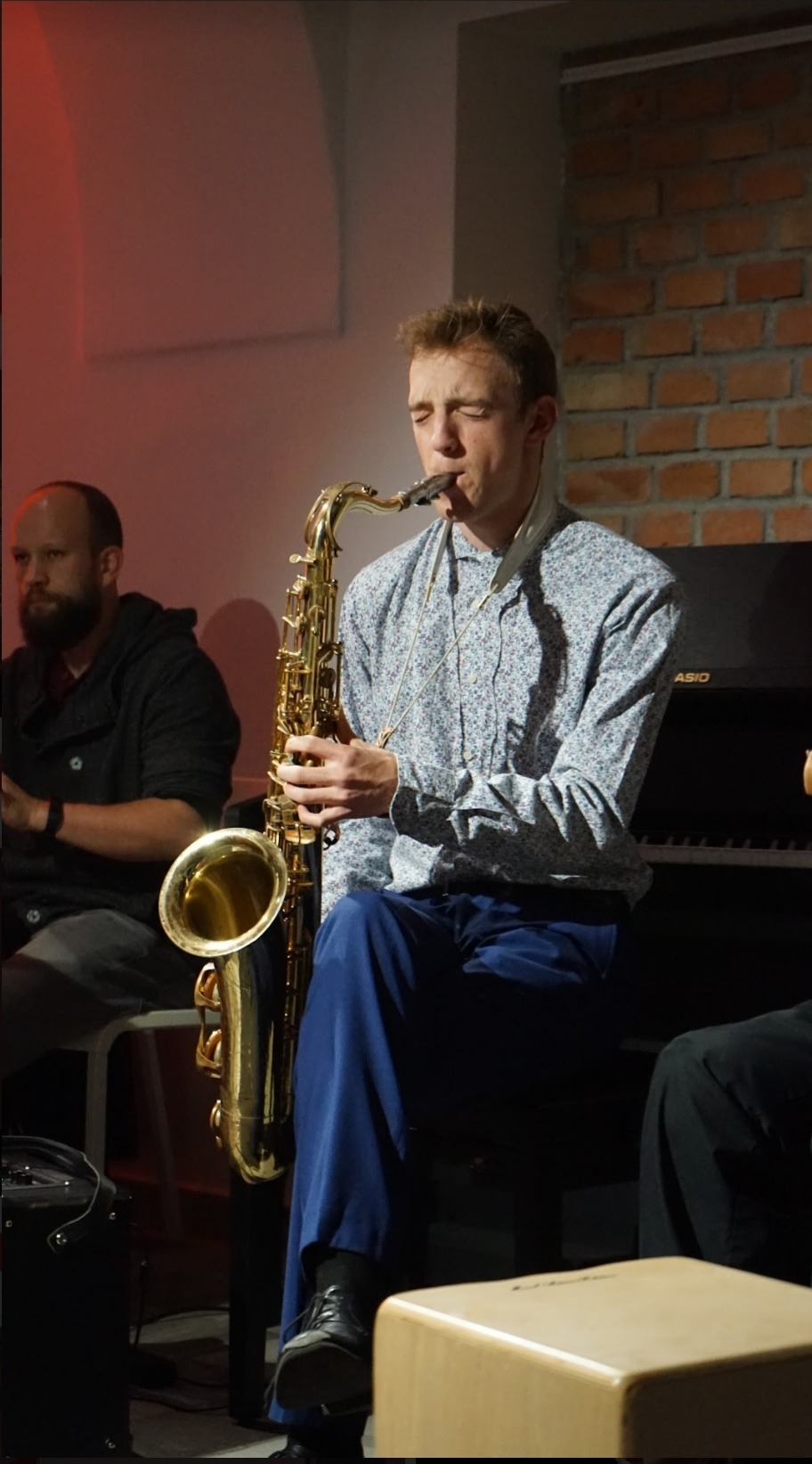 Saksofonista w kwiecistej koszuli gra na saksofonie tenorowym podczas występu na żywo; w tle widoczny perkusista i pianino.