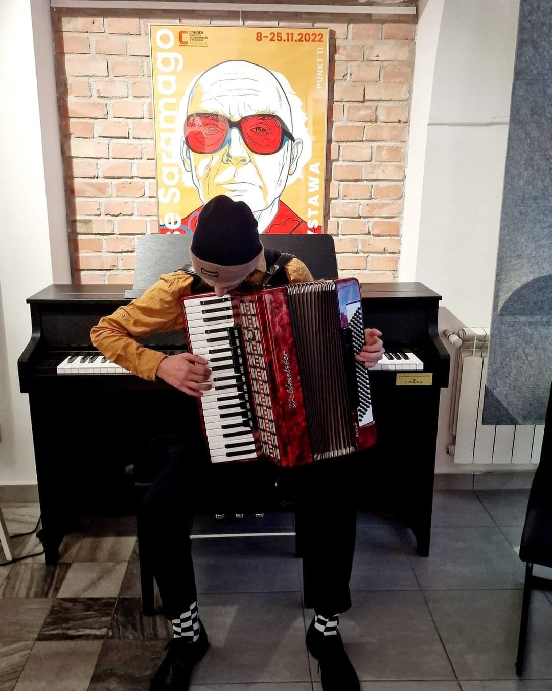 Młody mężczyzna w czapce gra na akordeonie marki Weltmeister, siedząc przed pianinem; w tle plakat z wizerunkiem osoby w czerwonych okularach na ceglanej ścianie.
