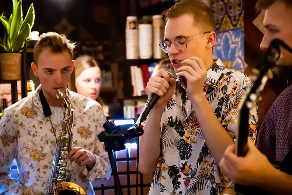 Młodzi muzycy w kwiecistych koszulach: saksofonista, wokalista w okularach z mikrofonem i gitarzysta podczas występu na tle regałów z książkami i roślinami.