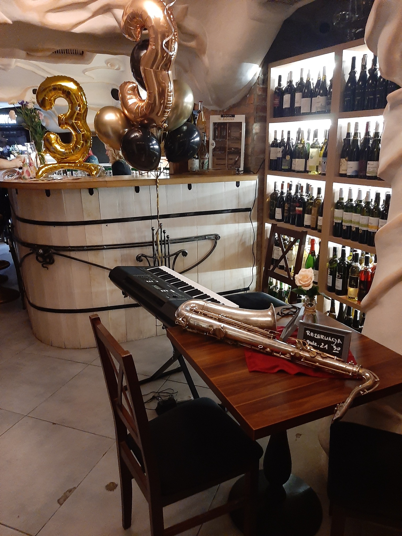 Saksofon tenorowy i keyboard na stoliku w restauracji, w tle dekoracja balonowa w kształcie cyfry 3 i półki z winami.