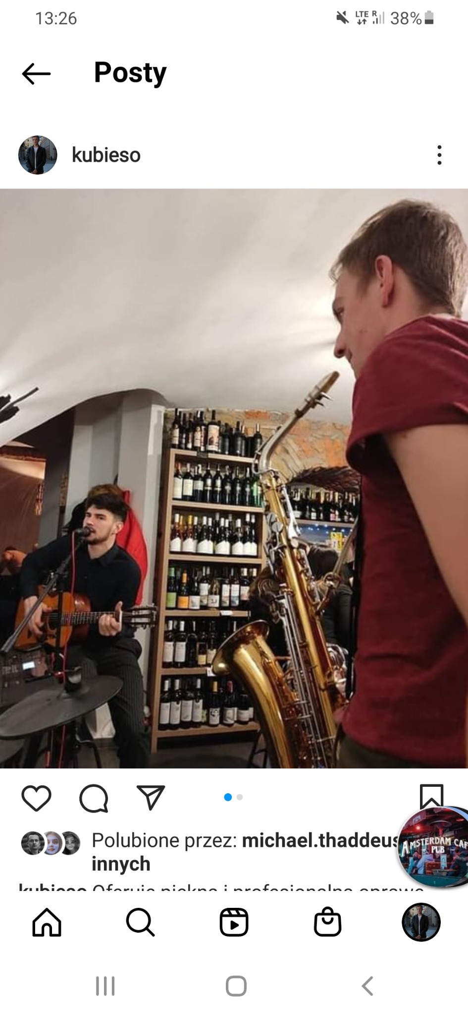 Występujący muzyk grający na gitarze i śpiewający do mikrofonu, obok saksofon, półki z butelkami w tle.