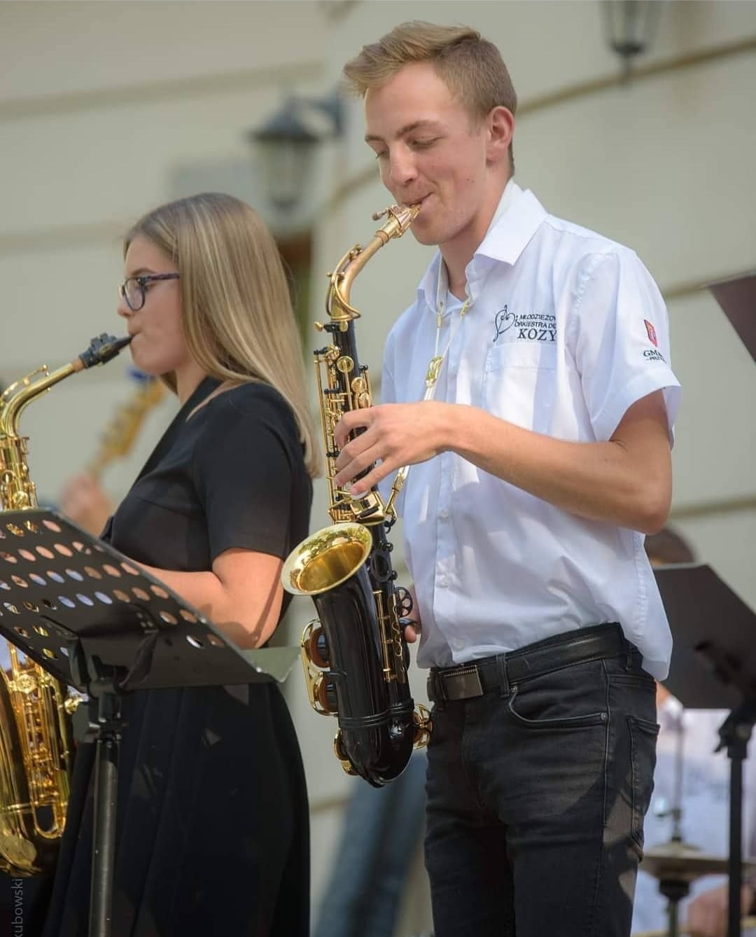 Dwoje młodych ludzi gra na saksofonach podczas występu plenerowego; mężczyzna w białej koszuli z logo orkiestry dętej.