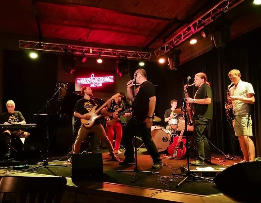 Zespół muzyczny grający jazz na scenie klubu z czerwonym oświetleniem, widoczni: wokalista, dwóch saksofonistów, gitarzysta basowy, gitarzysta, perkusista i pianista.