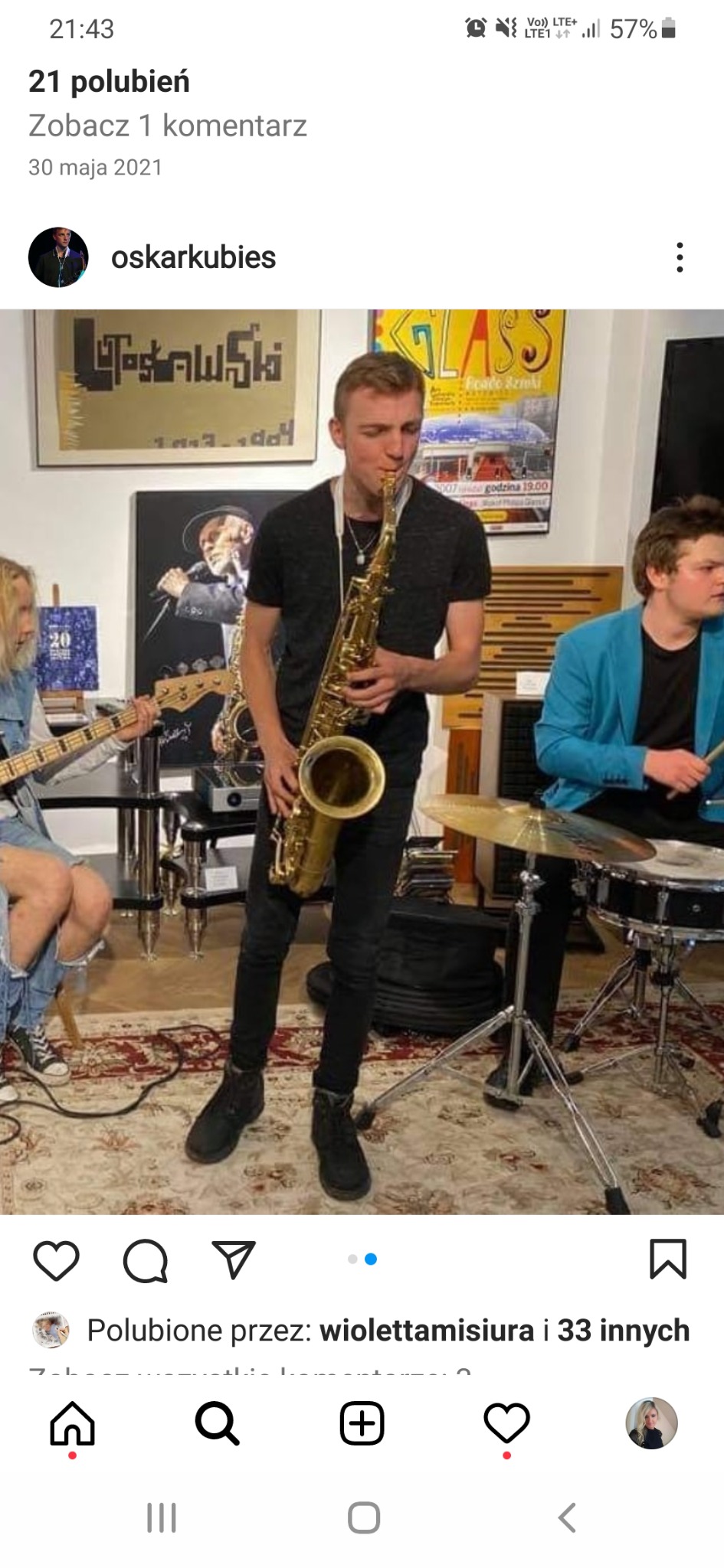 Saksofonista w czarnym ubraniu gra na saksofonie na tle zespołu muzycznego w pomieszczeniu z plakatami na ścianach i wzorzystym dywanem.