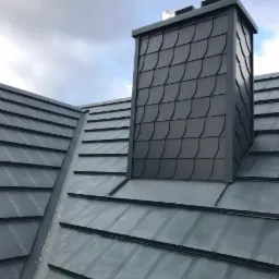 Nowoczesny dach z metalowej blachy w kolorze grafitowym, z kominem obłożonym łupkiem w kształcie rybiej łuski, sfotografowany pod pochmurnym niebem.