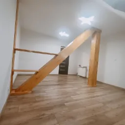 Wykończone wnętrze poddasza z drewnianą konstrukcją schodów i słupem podtrzymującym, panele podłogowe ułożone w jodełkę, białe ściany i sufit, widoczne grzejniki.