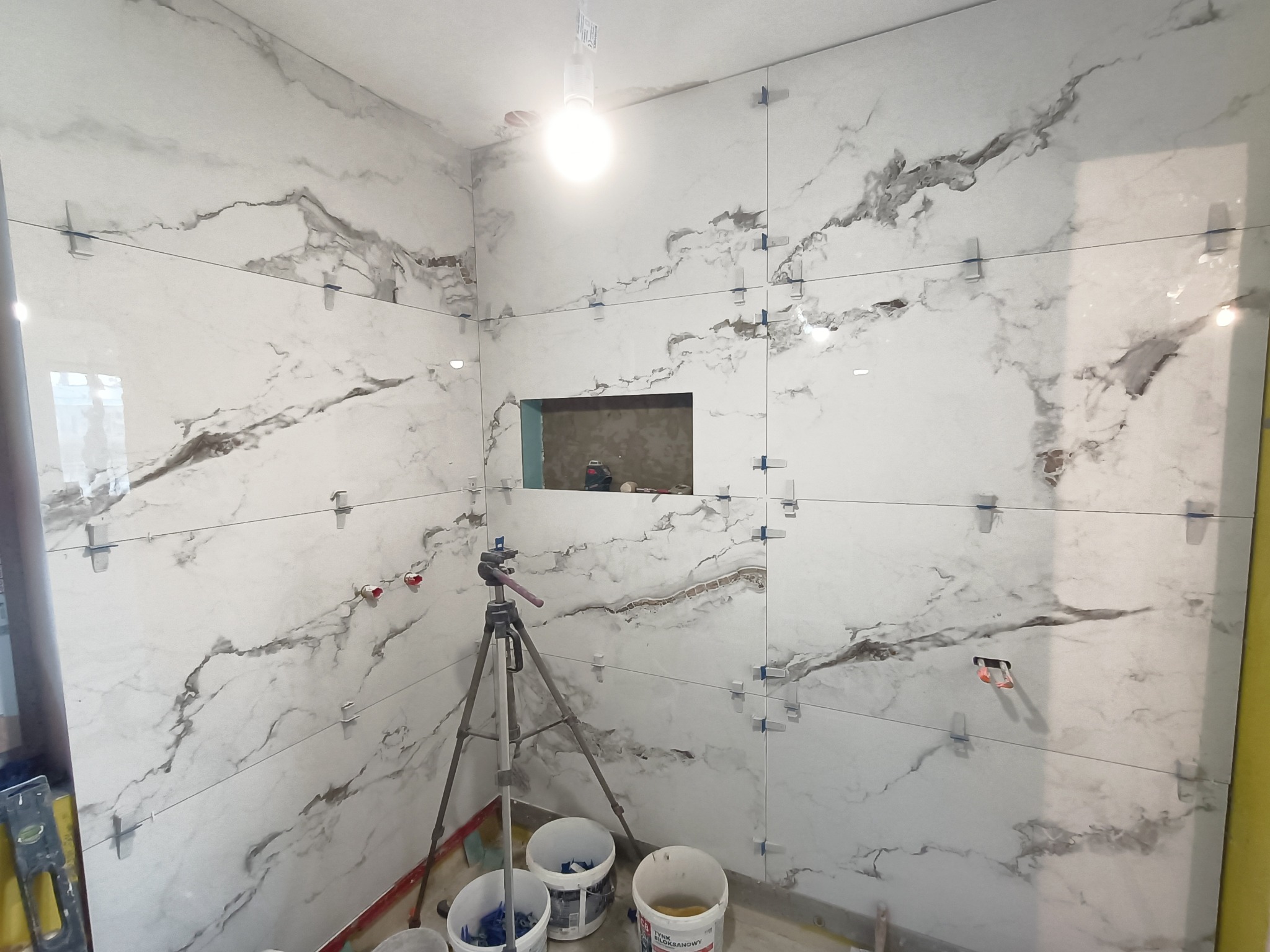 Ściany łazienki w trakcie układania płytek imitujących marmur, z użyciem klipsów poziomujących, widoczny otwór na instalację i statyw fotograficzny w rogu pomieszczenia.