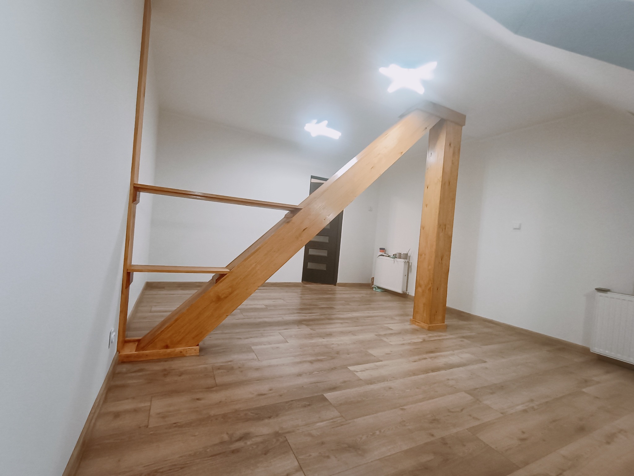 Wykończone wnętrze poddasza z drewnianą konstrukcją schodów i słupem podtrzymującym, panele podłogowe ułożone w jodełkę, białe ściany i sufit, widoczne grzejniki.