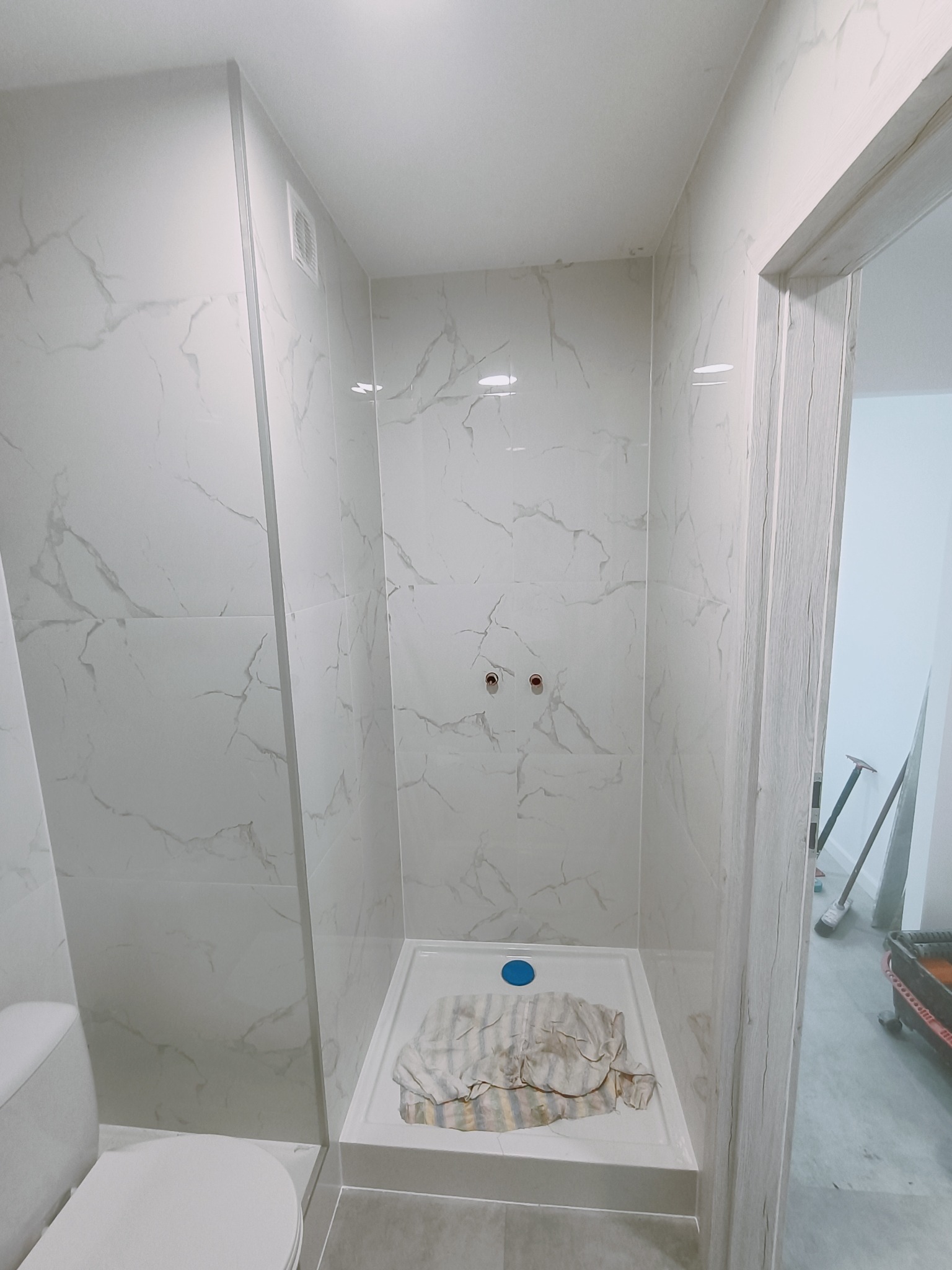 Wykończona kabina prysznicowa z białymi płytkami imitującymi marmur, widoczne przyłącza wodne, brodzik z niebieskim odpływem i leżący na nim ręcznik, obok toaleta i fragment otwartego wejścia...