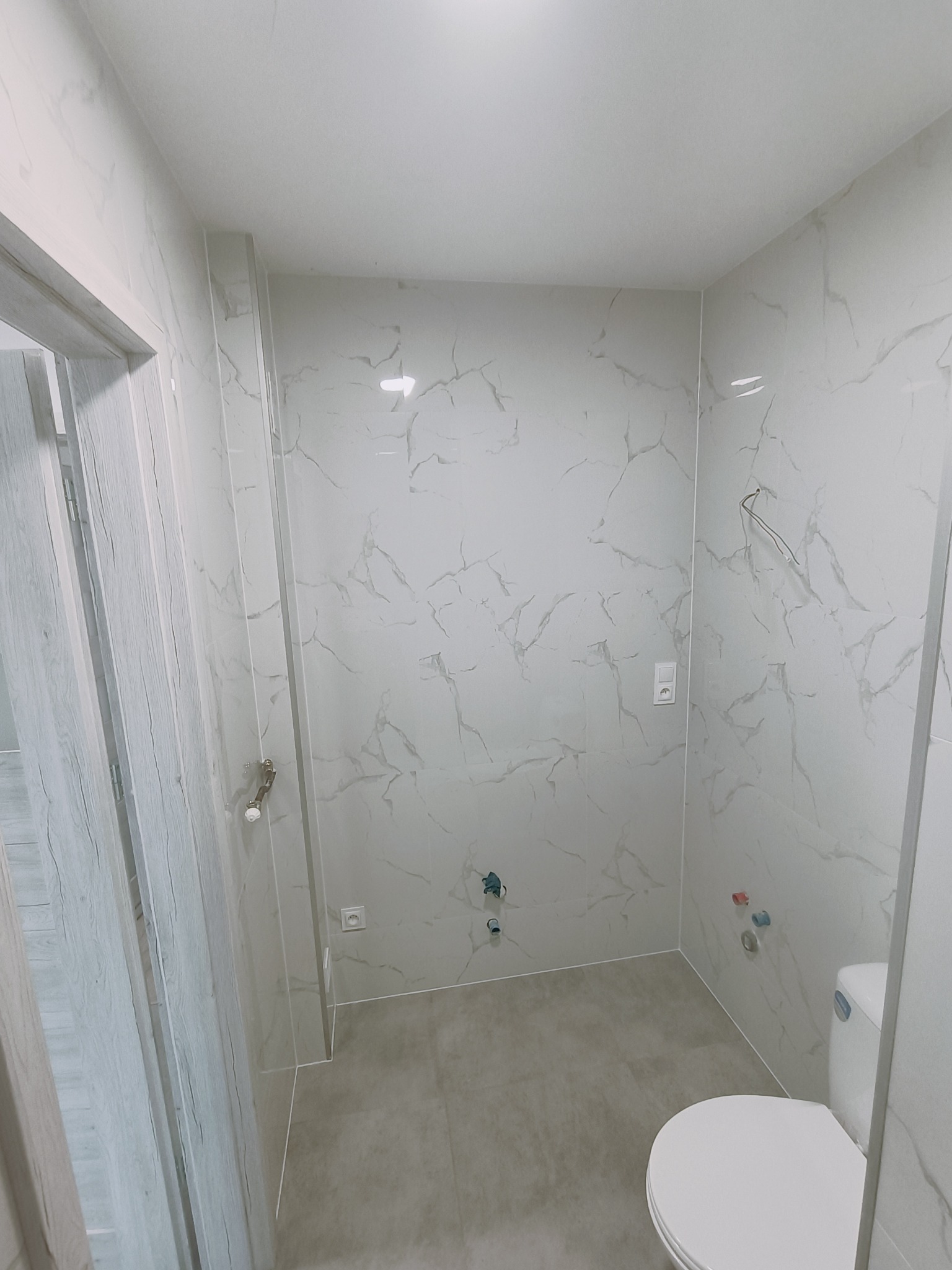 Wykończona łazienka z białymi płytkami imitującymi marmur na ścianach i podłodze, widoczne przyłącza wodne i częściowo zamontowana toaleta.