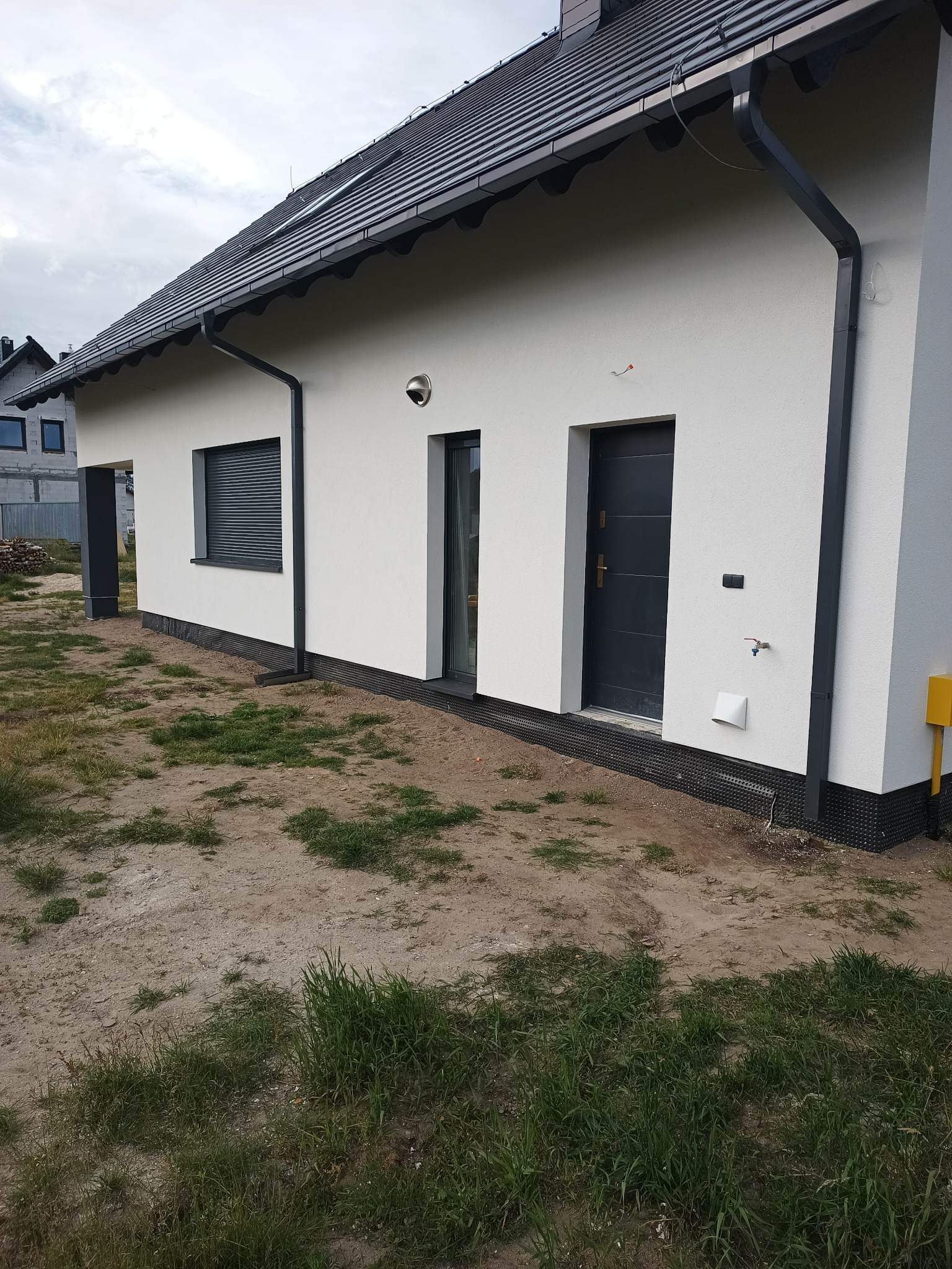 Nowo otynkowany, biały dom jednorodzinny z czarną stolarką okienną i drzwiami wejściowymi, widoczne orynnowanie, czarna listwa cokołowa i fragment żółtej skrzynki gazowej