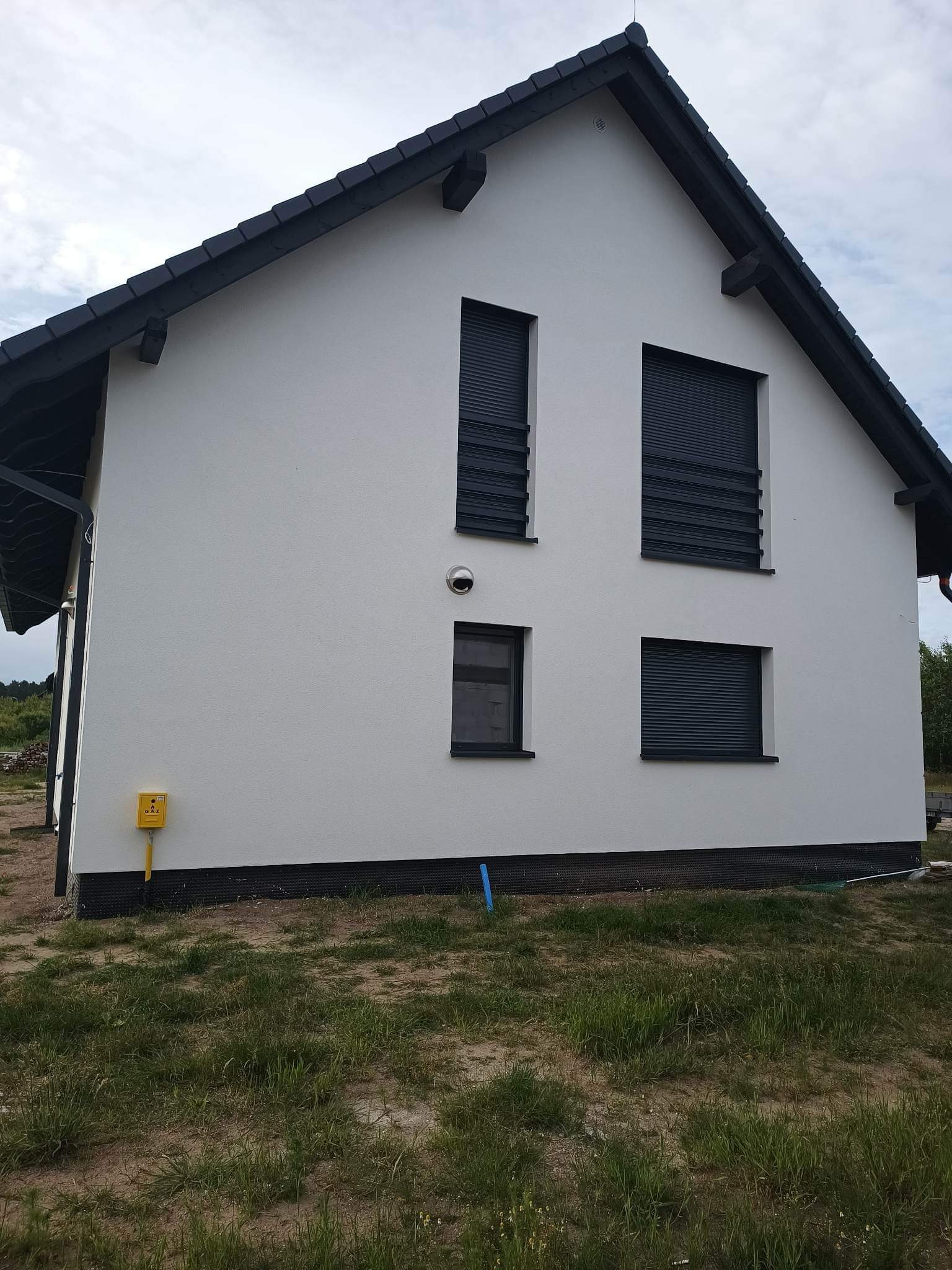 Nowoczesny dom jednorodzinny z białą elewacją i ciemnymi roletami zewnętrznymi, widok z boku na żółtą skrzynkę gazową i fragment trawnika.