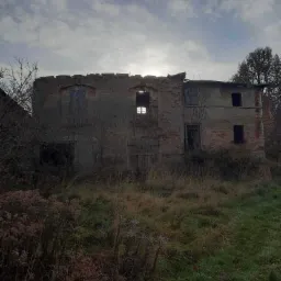 Zdjęcie zrzutu ekranu z komunikatora przedstawiające zrujnowany, opuszczony budynek z cegły i betonu, zarośnięty trawą i chwastami, z widocznymi oknami bez szyb i uszkodzonym dachem, w pochmurny...