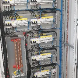 Otwarta szafa elektryczna z widocznymi bezpiecznikami, przewodami w kolorach czarnym i niebieskim, oraz szynami zbiorczymi. Żółte etykiety z oznaczeniami 'POLE 1.1', 'POLE 1.2', 'POLE 2.1', 'POLE 2...