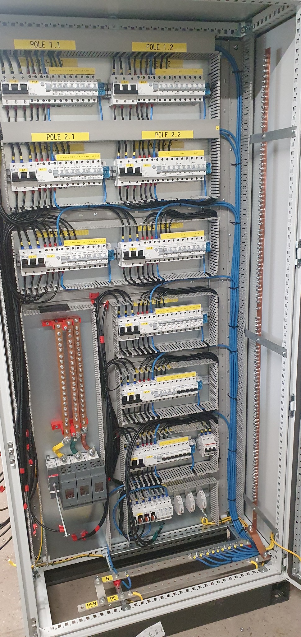 Otwarta szafa elektryczna z widocznymi bezpiecznikami, przewodami w kolorach czarnym i niebieskim, oraz szynami zbiorczymi. Żółte etykiety z oznaczeniami 'POLE 1.1', 'POLE 1.2', 'POLE 2.1', 'POLE 2...
