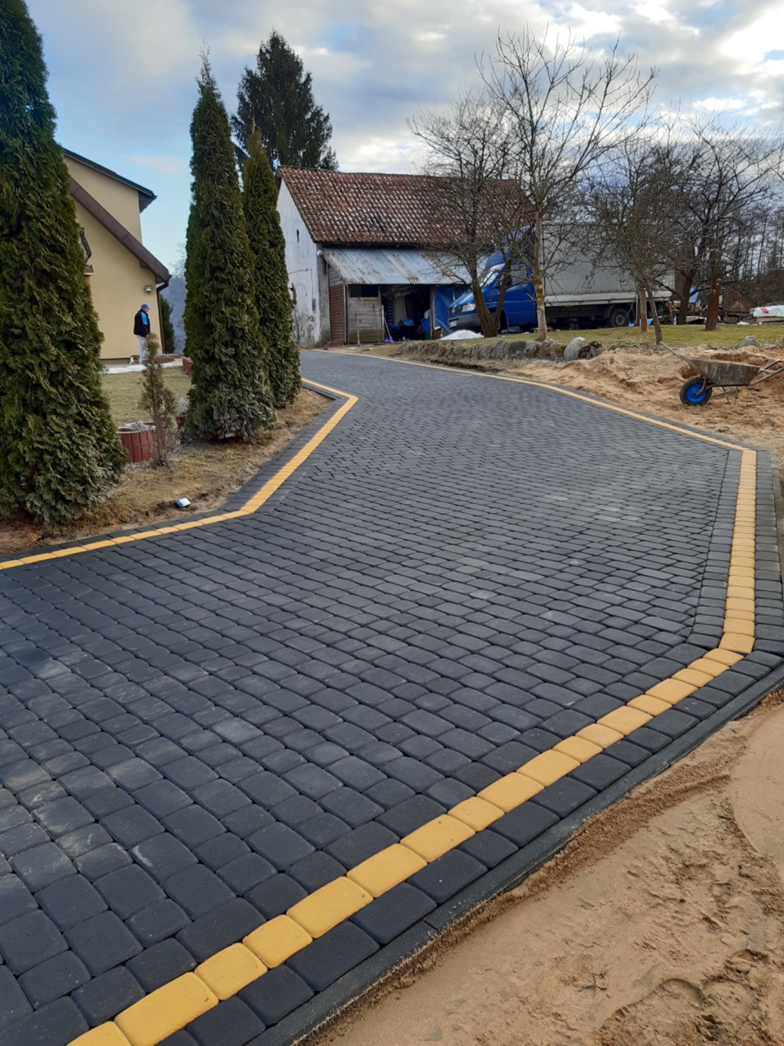 Nowo ułożona, wijąca się ścieżka z ciemnej kostki brukowej z żółtymi obrzeżami w Olsztynie, prowadząca w kierunku zabudowań gospodarczych i domu, widoczny wózek budowlany.