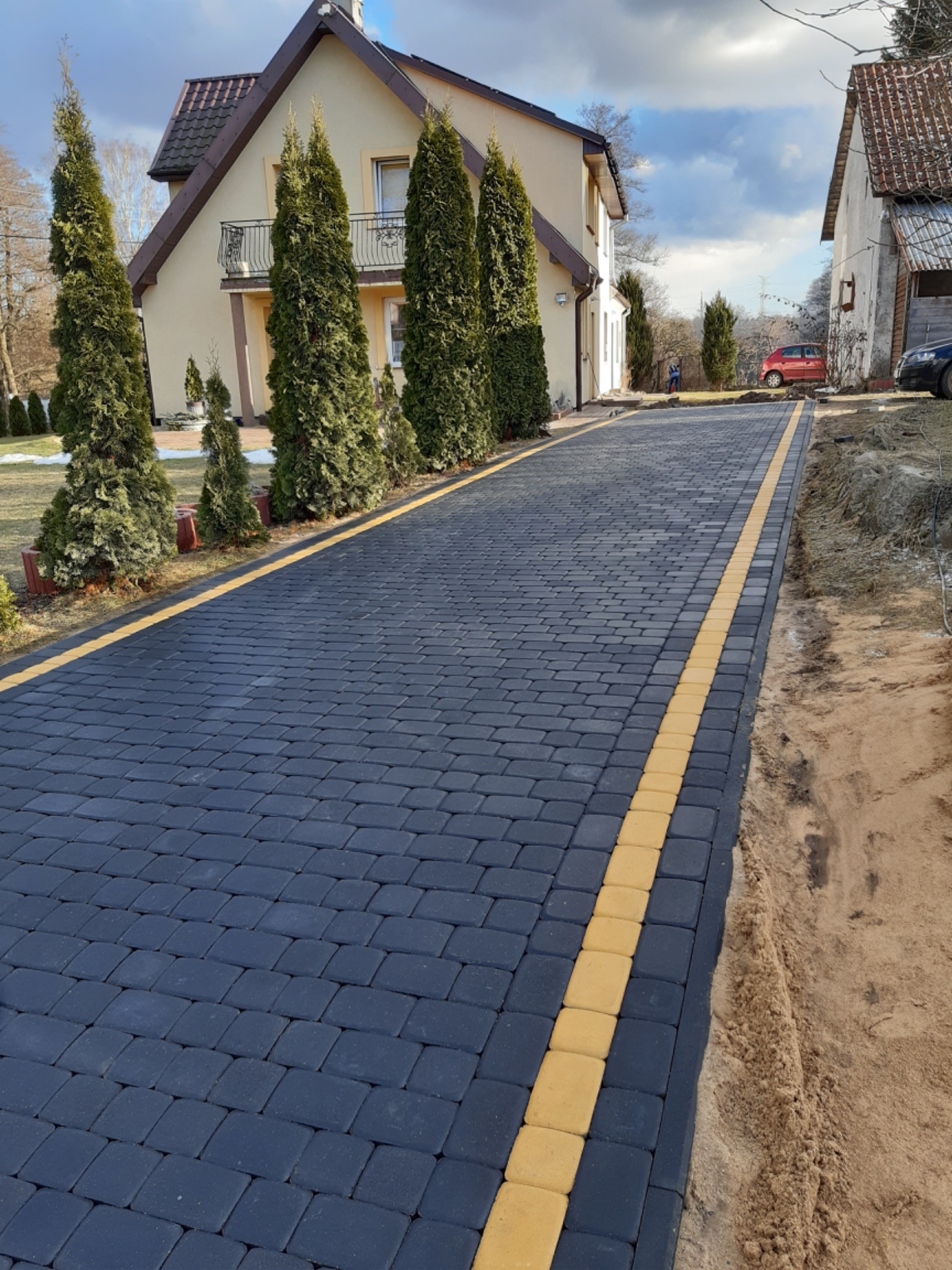 Ułożona, ciemnoszara kostka brukowa z żółtymi obrzeżami prowadząca do domu z balkonem, widok z perspektywy chodnika, świeżo obsadzone tuje po bokach.