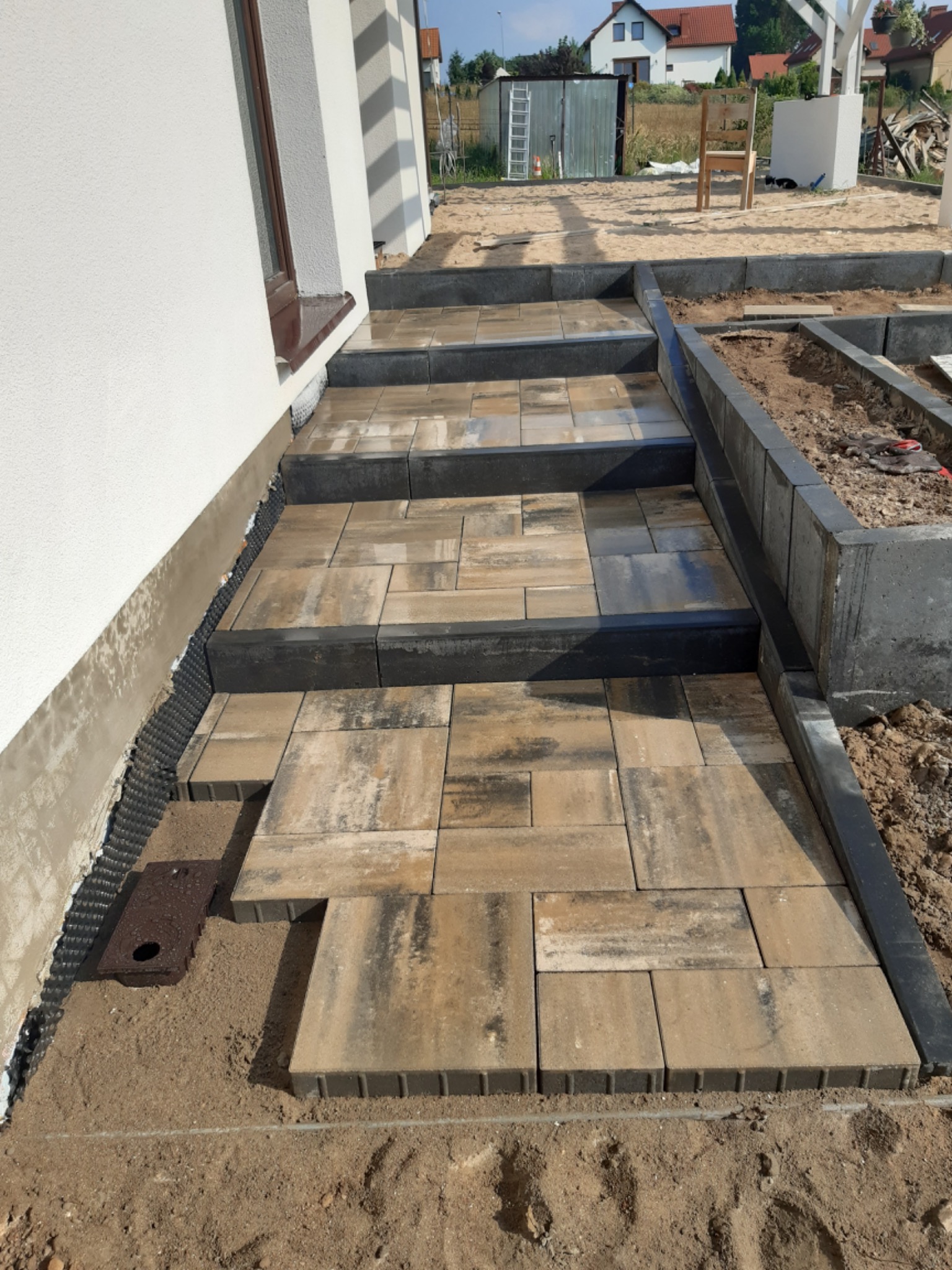 Schody wejściowe z kostki brukowej w trakcie układania, widoczne niedokończone obrzeża i elementy drenażowe przy ścianie budynku.