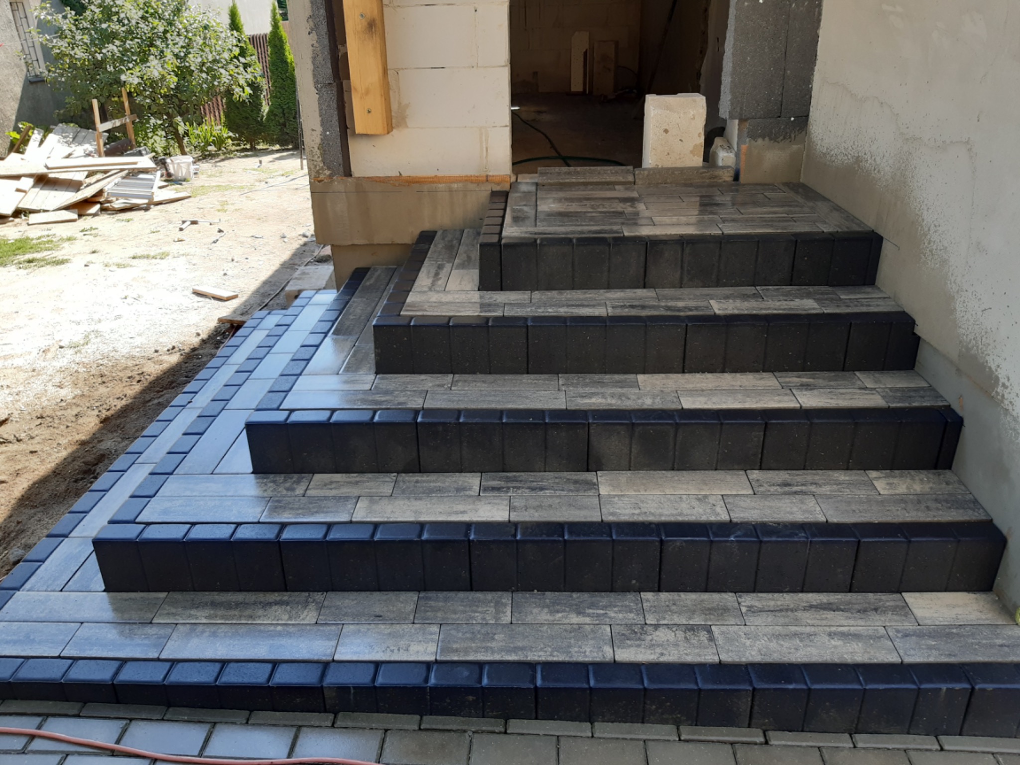 Schody wejściowe wyłożone kostką brukową w odcieniach szarości i grafitu, prowadzące do surowego stanu budynku, z widocznymi materiałami budowlanymi w tle.