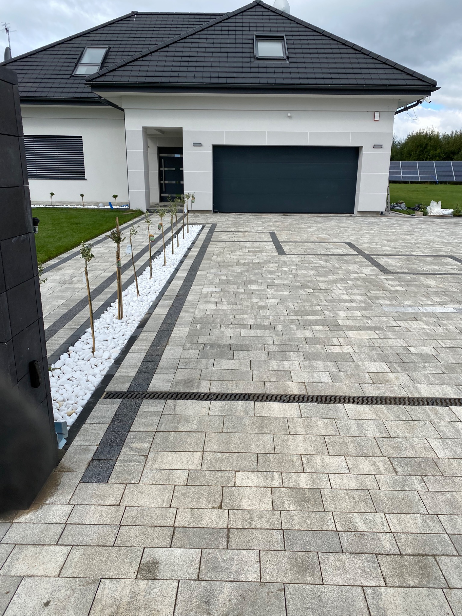 Ułożona szara kostka brukowa przed nowoczesnym domem z czarną bramą garażową i rzędem małych drzewek oddzielonych białymi kamieniami, widoczna krata odpływowa.