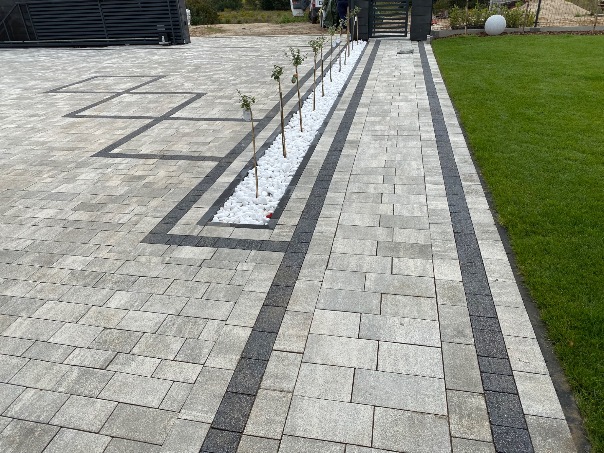 Ułożona kostka brukowa w odcieniach szarości, z dekoracyjnym pasem z białych kamieni i rzędem młodych drzewek, otoczona świeżym trawnikiem i nowoczesną bramą.