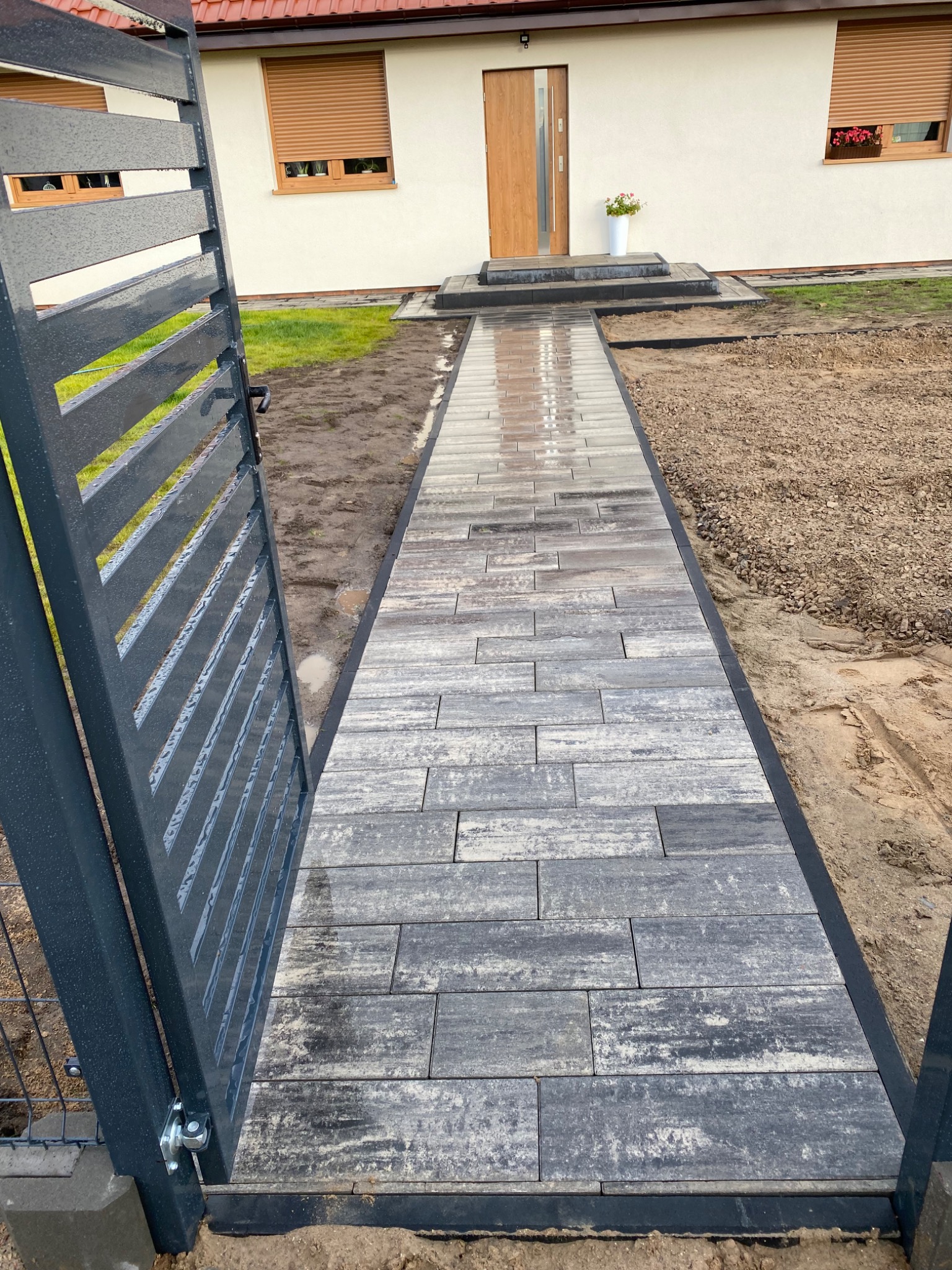 Nowo ułożona, wilgotna ścieżka z szarej kostki brukowej prowadząca od metalowej bramy w kierunku wejścia do domu z beżową elewacją i brązowymi roletami.