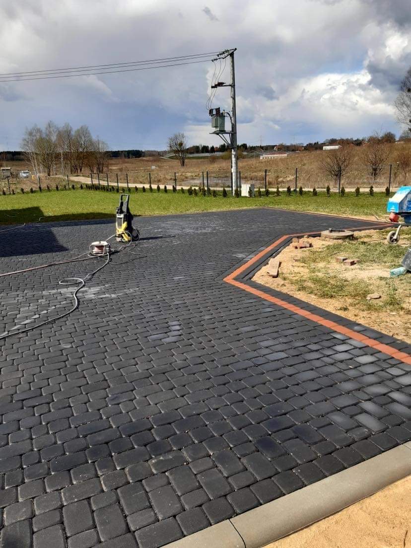 Ułożona, ciemnoszara kostka brukowa z pomarańczowym obramowaniem, widoczna myjka ciśnieniowa i częściowo przygotowany teren pod dalsze prace.