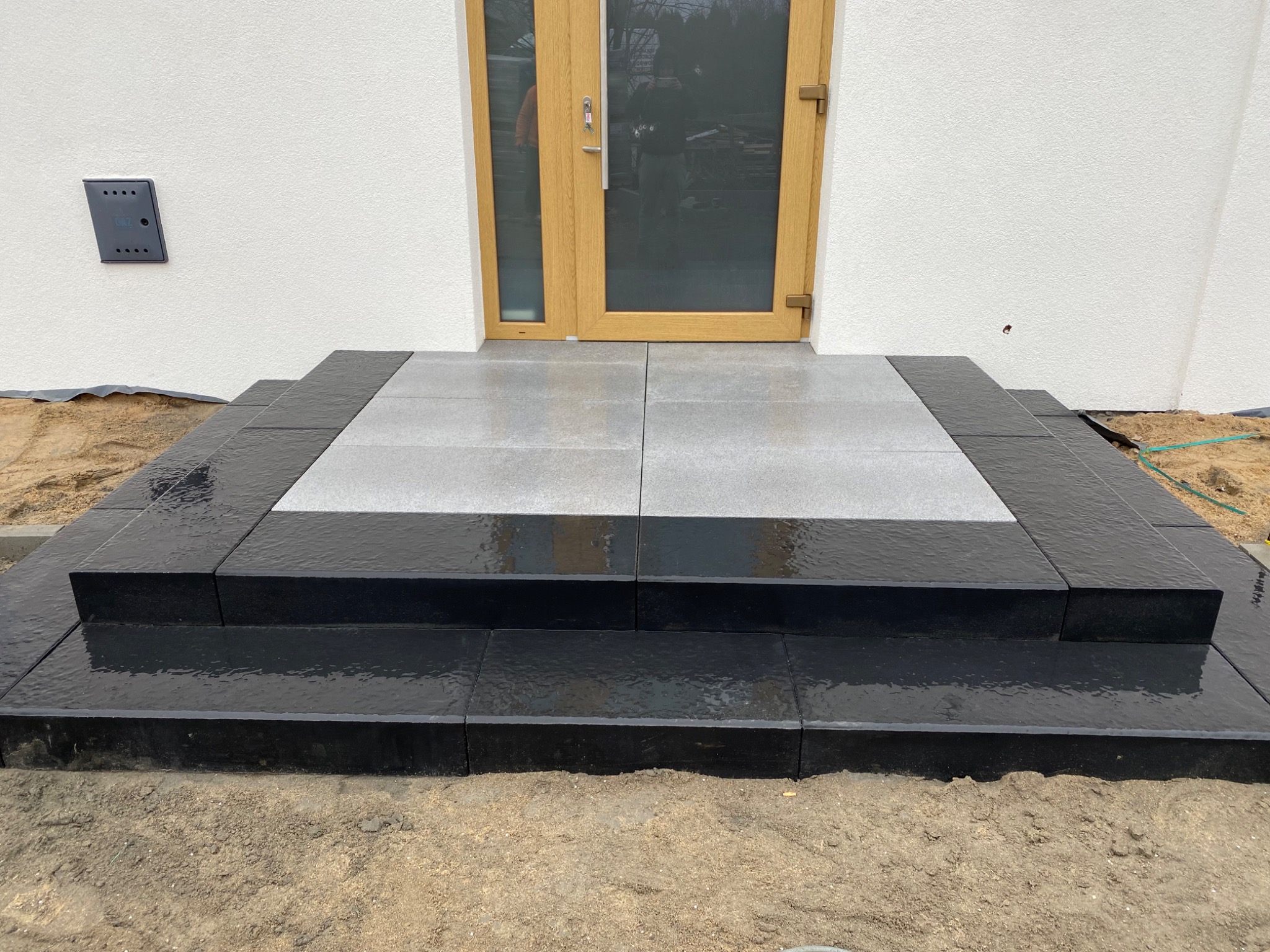 Nowe, dwukolorowe schody wejściowe z czarnej i szarej kostki brukowej prowadzące do drewnianych drzwi wejściowych w jasnym budynku, widoczne ślady wilgoci na powierzchni kostki.