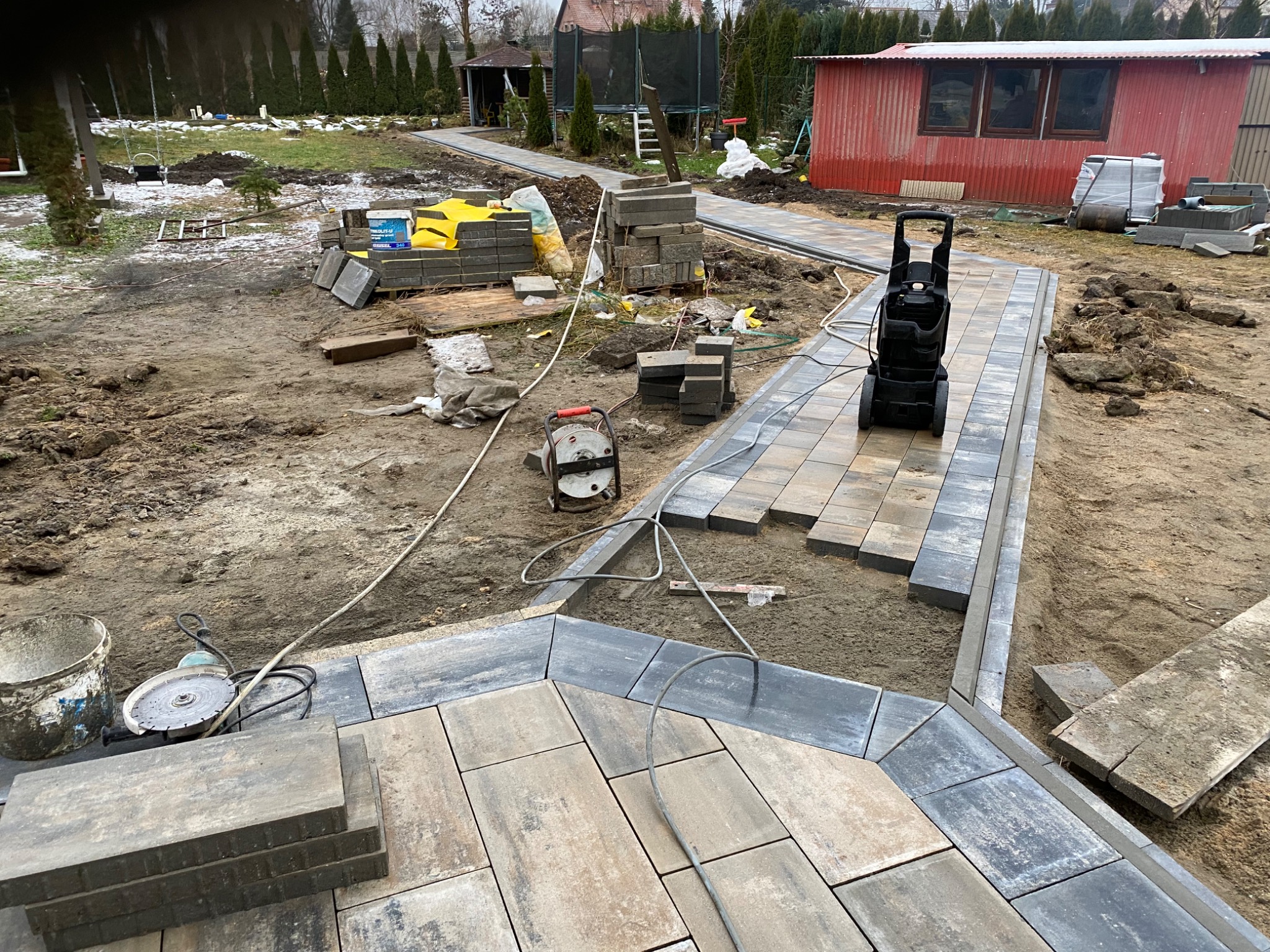 Fragment ścieżki z kostki brukowej w trakcie układania, widoczna przecinarka do kostki, palety z kostką w tle, teren przygotowany pod dalsze prace.