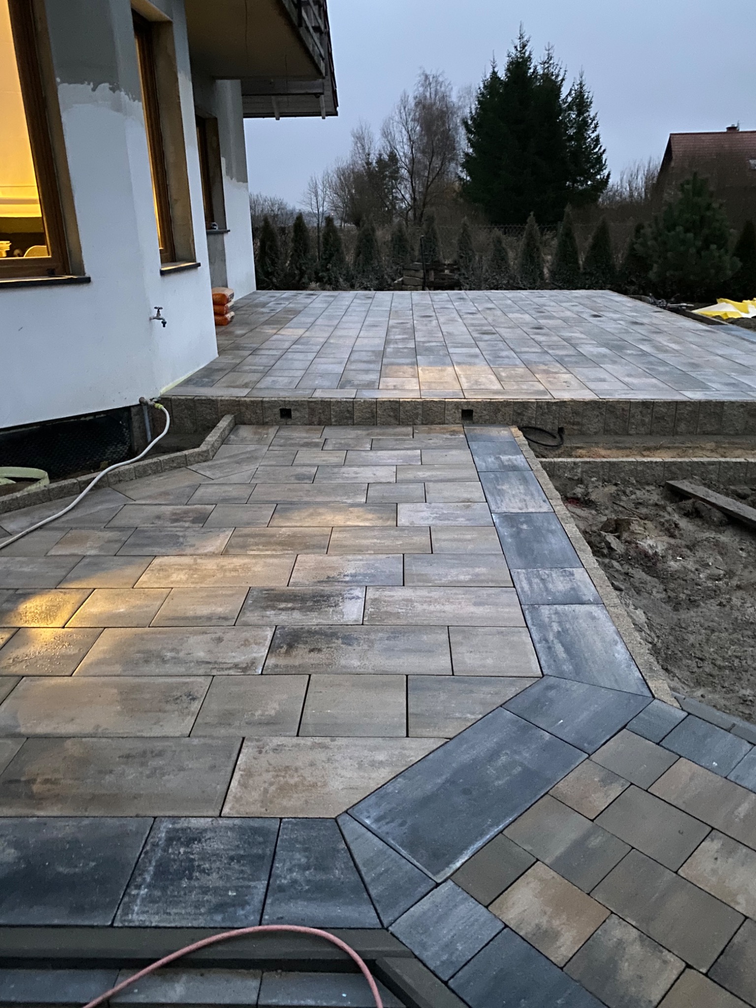 Ułożona kostka brukowa na podwórku, z widocznymi różnicami kolorystycznymi kostek i obrzeżami w odcieniach szarości, widok na dom i ogród w tle, częściowo ukończony fragment z odkrytą ziemią.