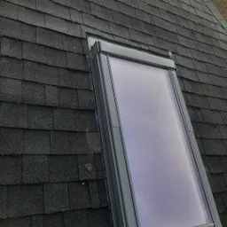 Szczegółowe ujęcie okna dachowego osadzonego w pokryciu z czarnej dachówki bitumicznej. Widoczne ślady wilgoci na szybie i drobne zabrudzenia na dachówkach.