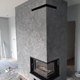Wykończony kominek narożny z szarą, dekoracyjną strukturą na ścianie, czarne elementy wykończeniowe, zabezpieczona podłoga folią malarską, wnętrze w trakcie prac wykończeniowych.