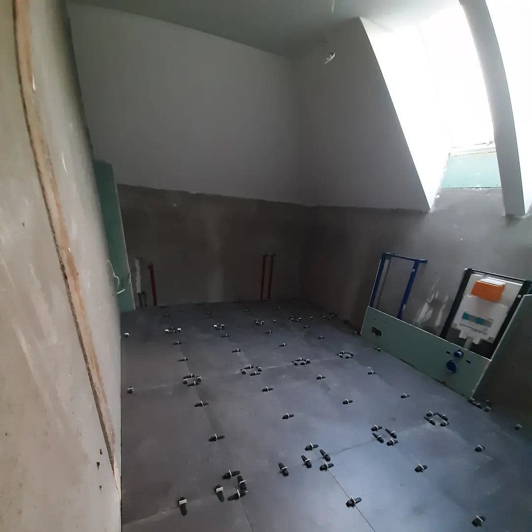 Łazienka w trakcie remontu: szare płytki podłogowe z plastikowymi krzyżykami dystansowymi, częściowo wykończone ściany, widoczne rury instalacji wodnej i stelaż podtynkowy WC.