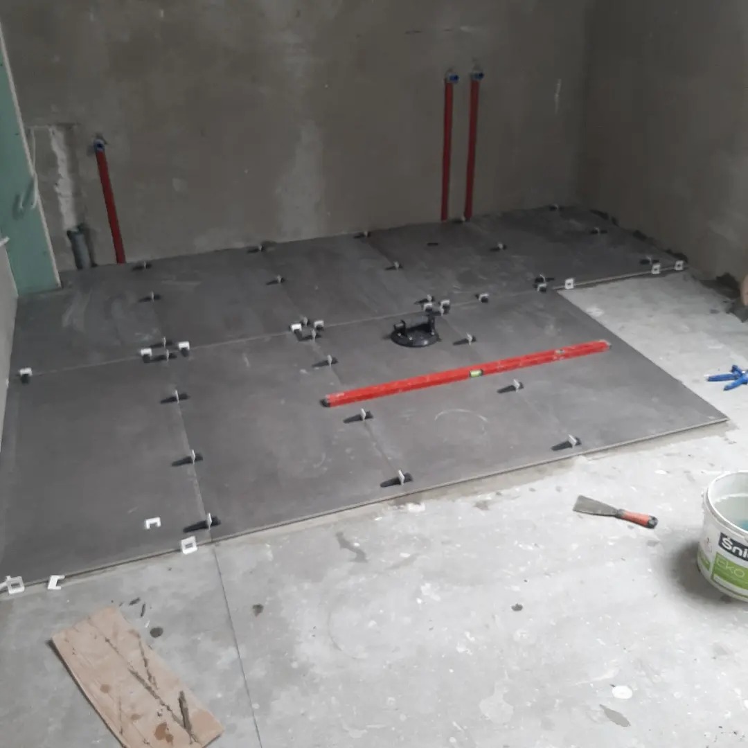 Układanie ciemnych płytek podłogowych w surowej łazience, widoczne krzyżyki dystansowe, poziomica, przyssawka do płytek i wiadro z zaprawą.