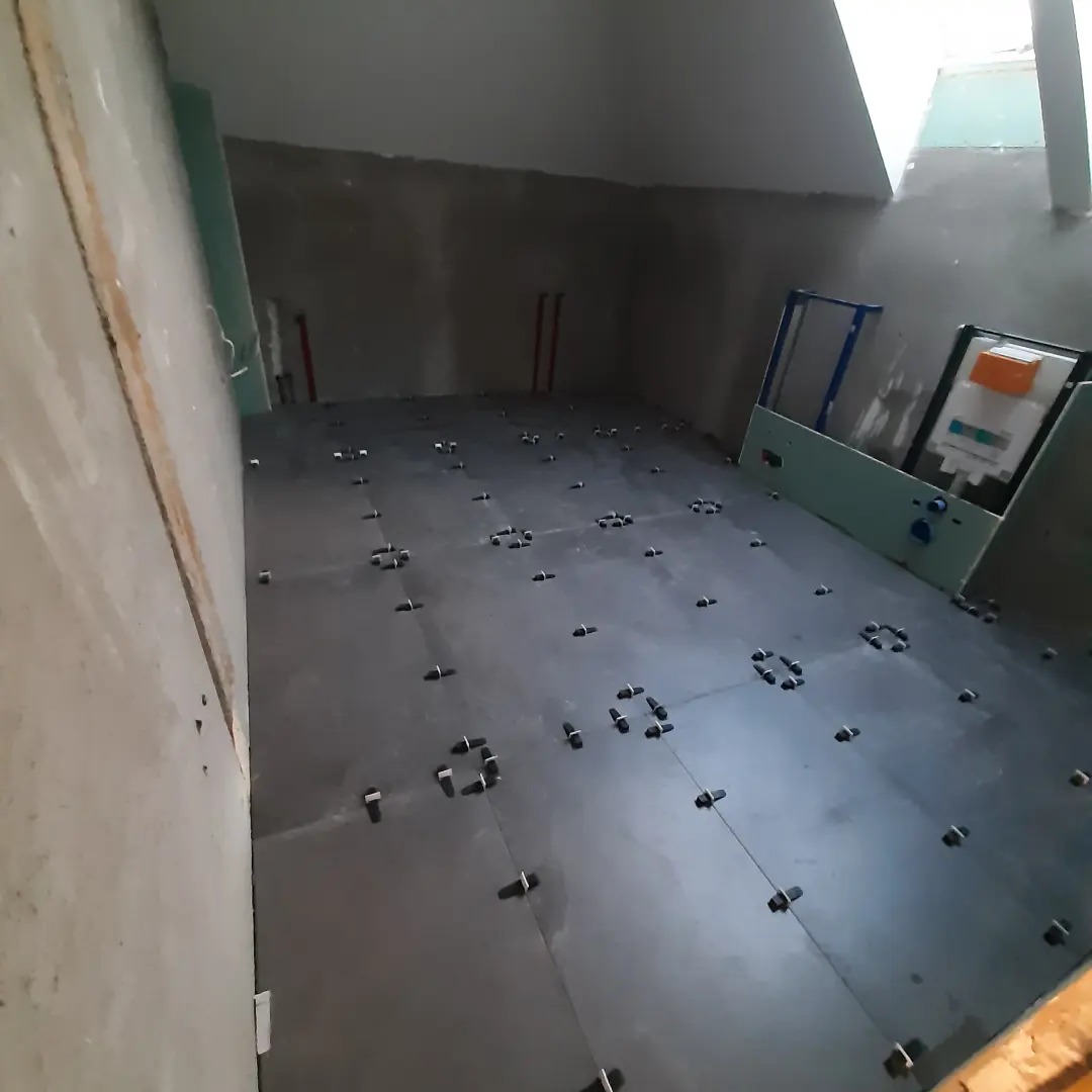 Łazienka w trakcie remontu: ciemne płytki podłogowe z plastikowymi krzyżykami dystansowymi, surowe ściany z instalacjami, zabudowa stelaża podtynkowego WC.