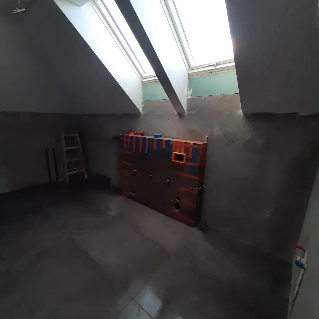 Łazienka na poddaszu w trakcie remontu: szare ściany, okno dachowe, zabudowa stelaża podtynkowego WC z kolorowymi oznaczeniami, widoczna drabina i przygotowane podłoże pod płytki.