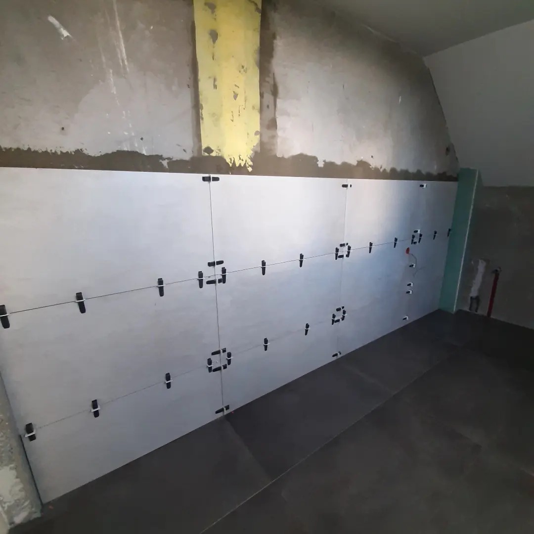 Świeżo ułożone, jasne płytki na ścianie w trakcie remontu łazienki, z użyciem krzyżyków dystansowych i sznurka do poziomowania, widoczne surowe ściany i podłoga w ciemnych płytkach.