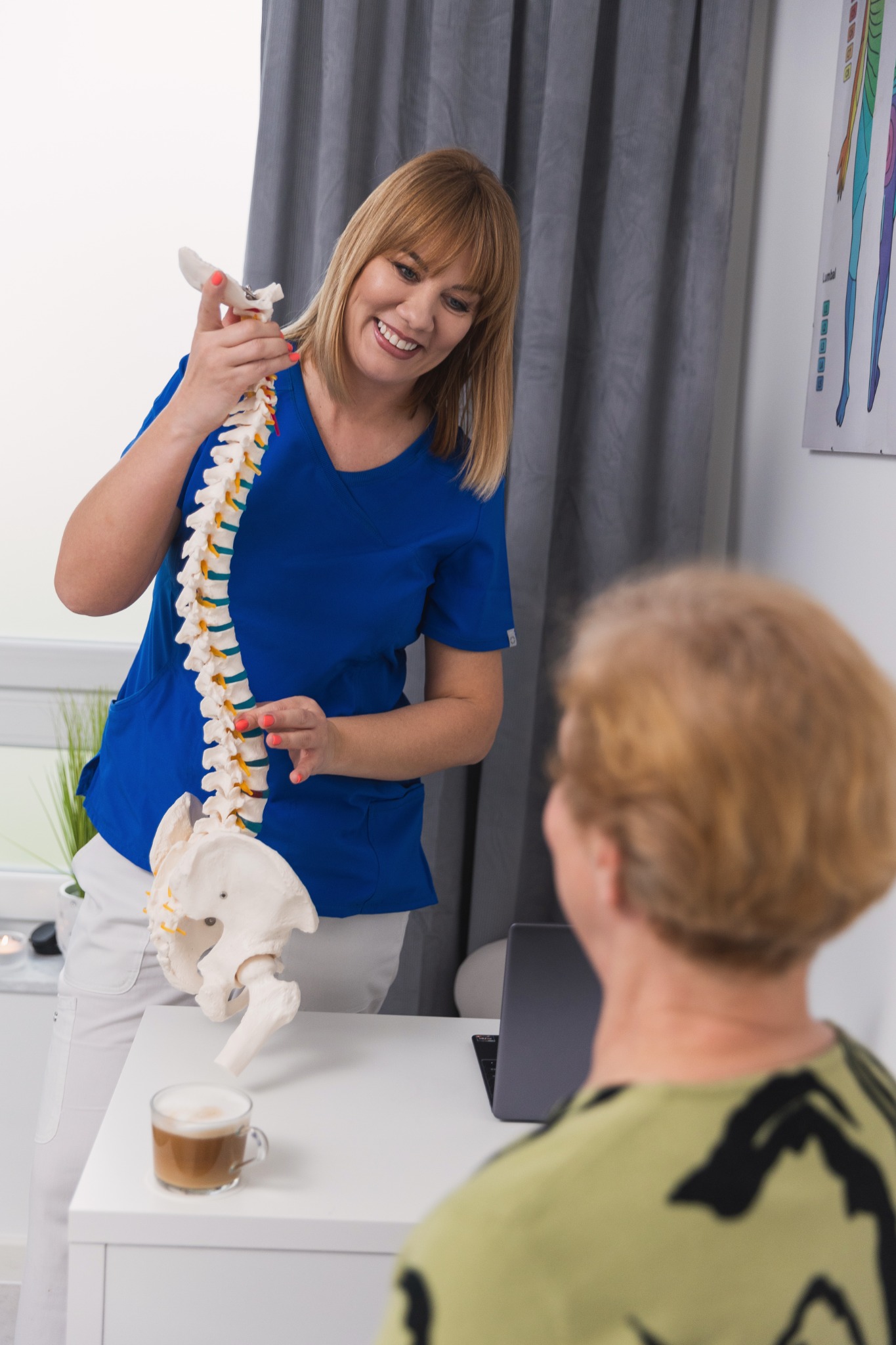 Kobieta w niebieskim uniformie demonstruje model kręgosłupa pacjentce w gabinecie, na biurku stoi kawa.