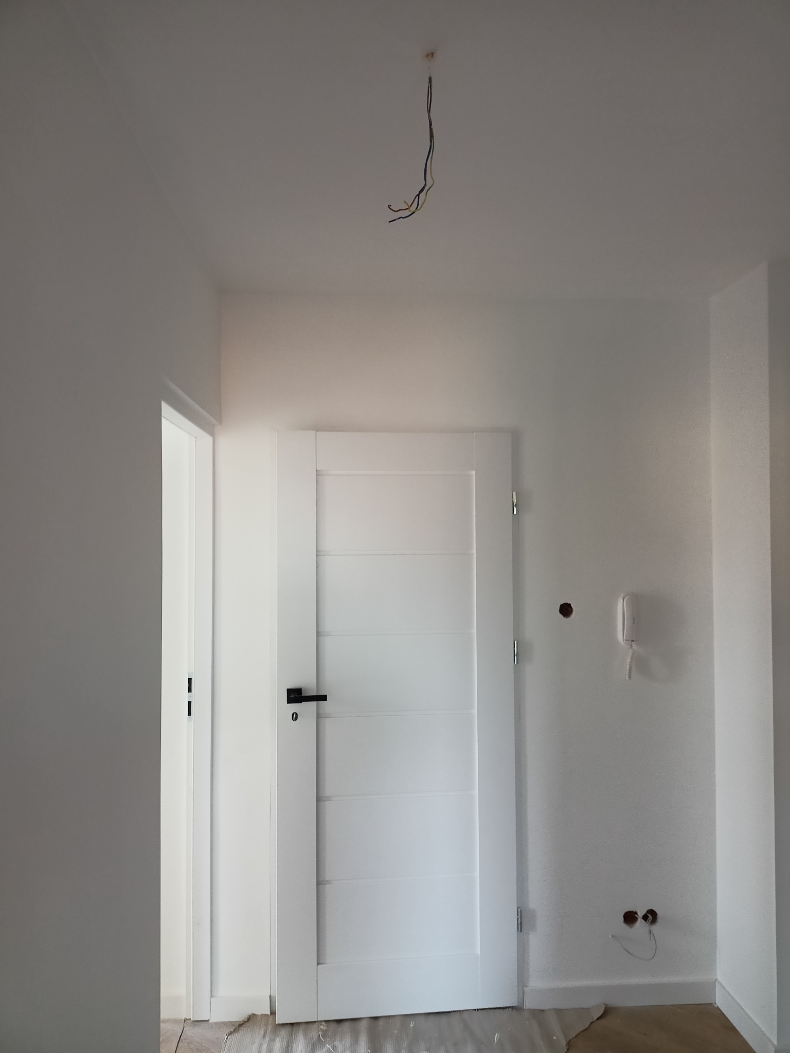 Białe wnętrze w trakcie malowania, widoczne świeżo pomalowane ściany, białe drzwi z czarną klamką, wystające kable z sufitu, gniazdka elektryczne bez osprzętu i niezabezpieczona podłoga.