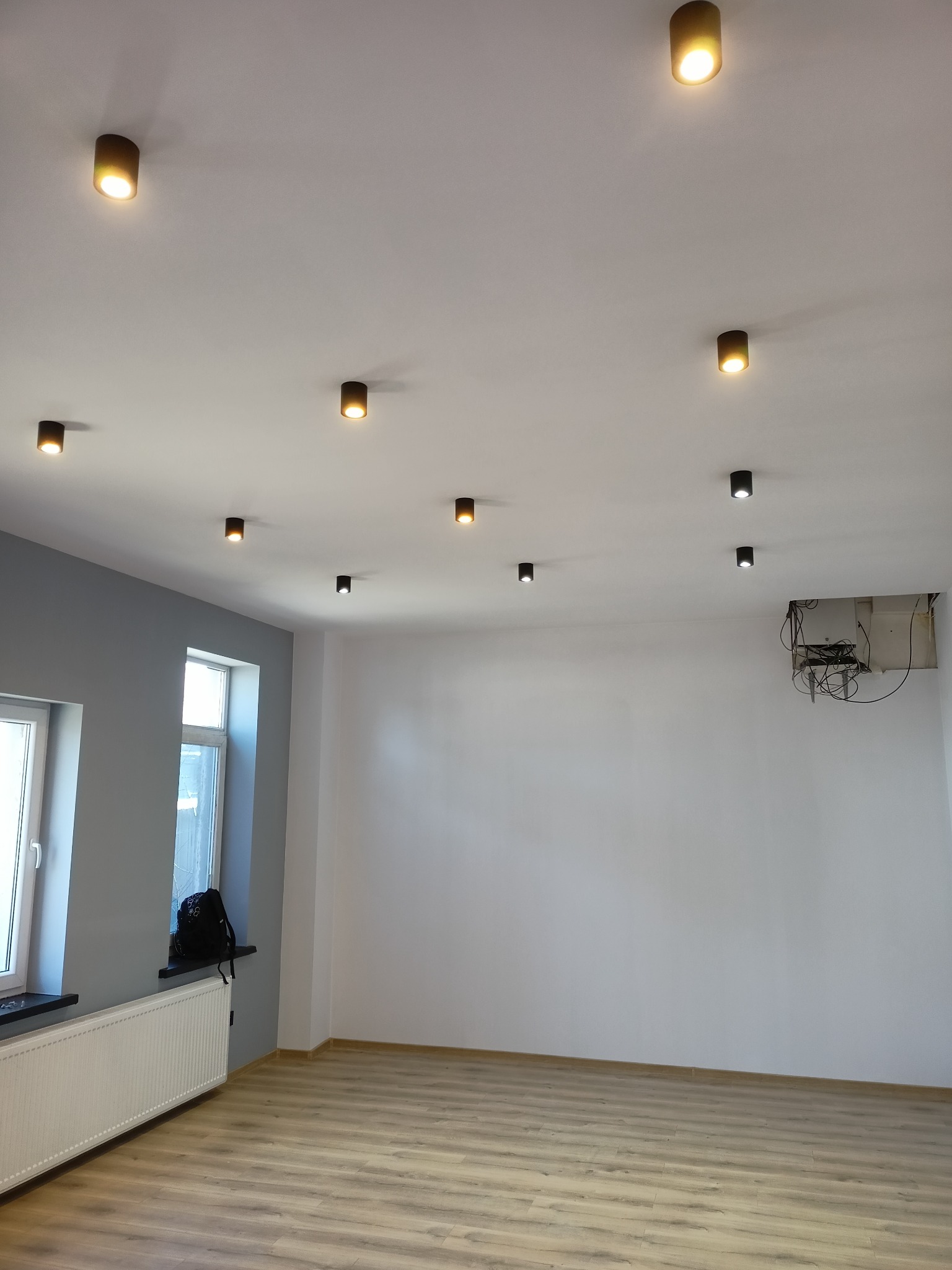 Pomalowane na biało pomieszczenie z panelami podłogowymi, grzejnikiem pod oknem i punktowym oświetleniem sufitowym. Widoczna instalacja elektryczna w suficie.