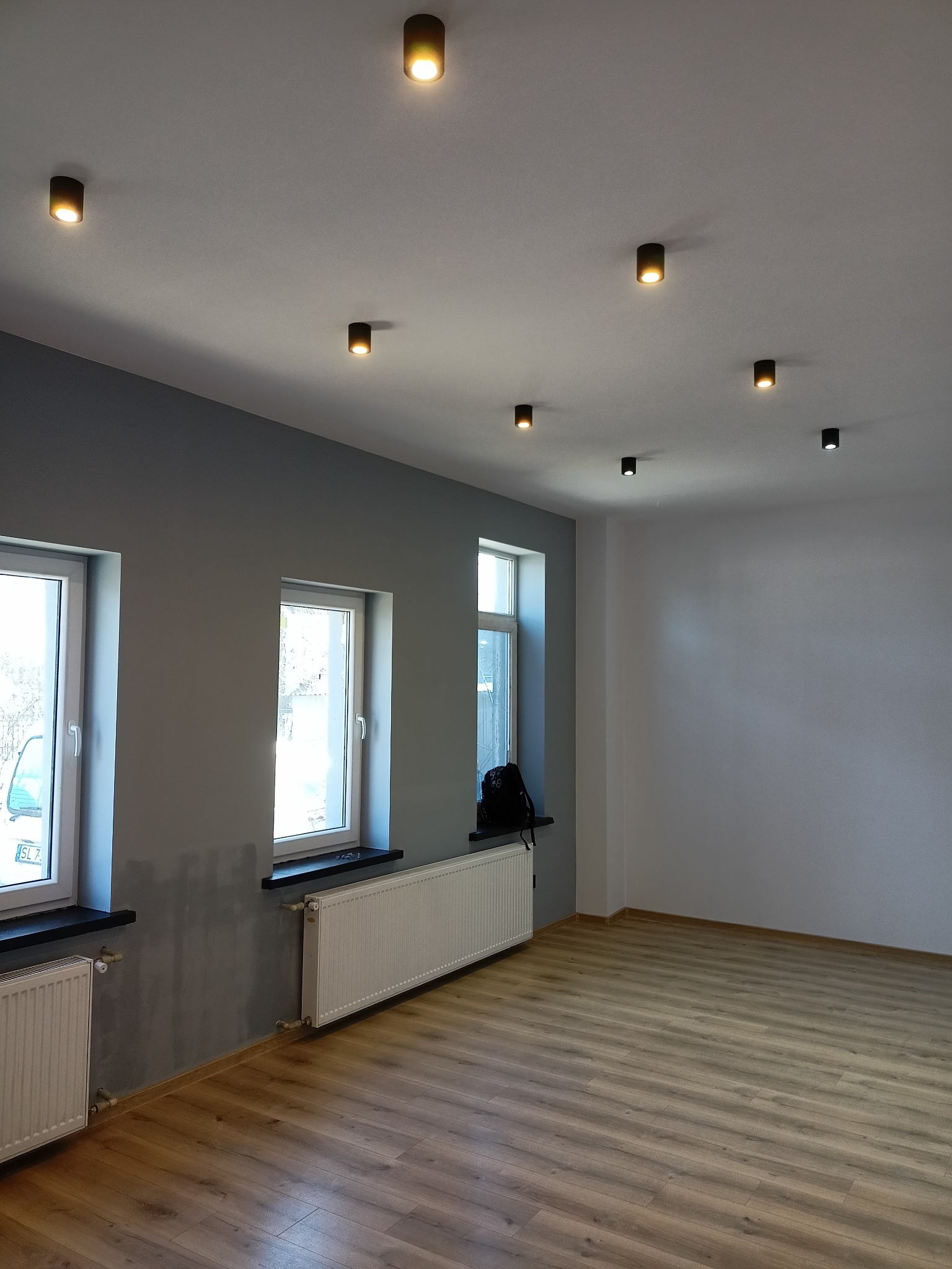 Wnętrze po remoncie: świeżo pomalowane szare ściany z widocznymi śladami po szpachlowaniu, drewnopodobna podłoga, białe grzejniki pod oknami i nowoczesne, punktowe oświetlenie sufitowe.
